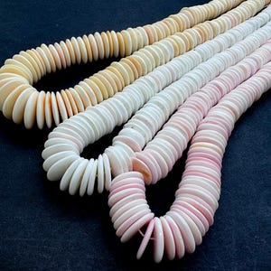 May include: Four strands of disc-shaped necklaces in varying shades of cream, white, and pink. The necklaces are arranged on a dark blue surface, showcasing the different colors and textures of the beads. The beads appear to be made of a natural material.