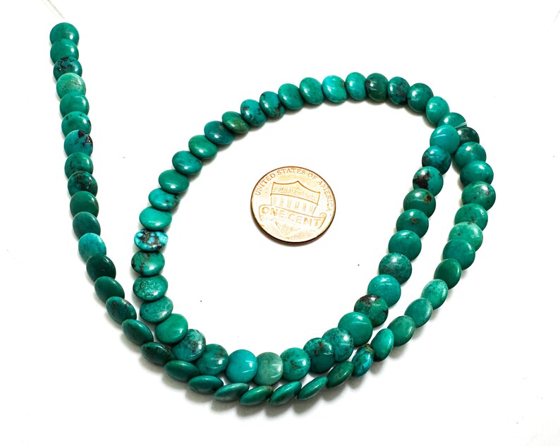 May include: A strand of round, flat turquoise beads with natural dark matrix veining, arranged in a loose loop. A copper-colored United States one-cent coin is placed in the center for scale, measuring approximately 0.75 inches in diameter.