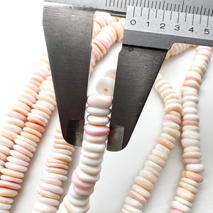 Op de afbeelding: Close-up van strengen roze en witte schelpkralen. Een Mitutoyo-schuifmaat meet de diameter van de kralen. De kralen zijn aan elkaar geregen, waardoor een ketting of armband ontstaat. De kralen hebben een platte, schijfvormige vorm.