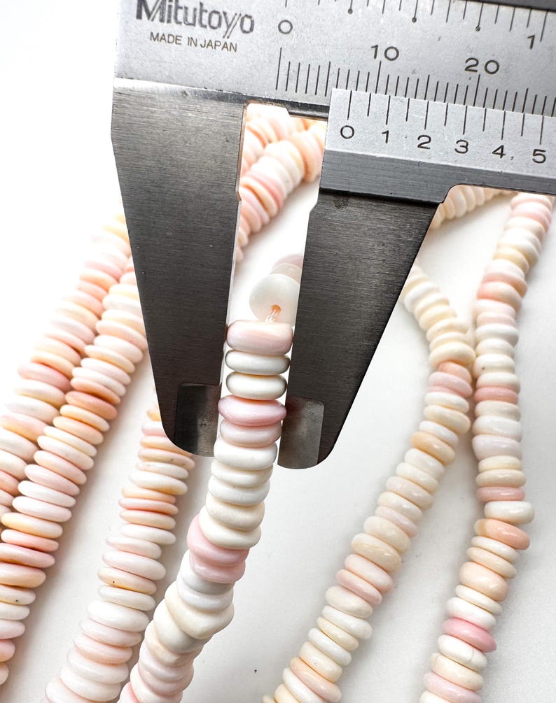 Op de afbeelding: Close-up van strengen roze en witte schelpkralen. Een Mitutoyo-schuifmaat meet de diameter van de kralen. De kralen zijn aan elkaar geregen, waardoor een ketting of armband ontstaat. De kralen hebben een platte, schijfvormige vorm.