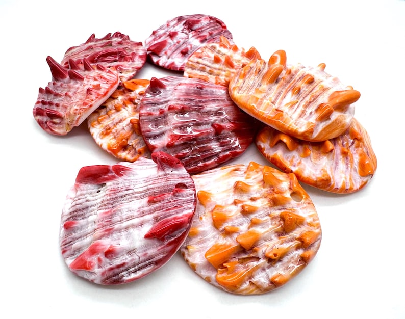 May include: A collection of decorative seashells in shades of red, orange, and white. The shells have a textured surface and a variety of shapes, including rounded and spiky designs. The shells are arranged on a white background.