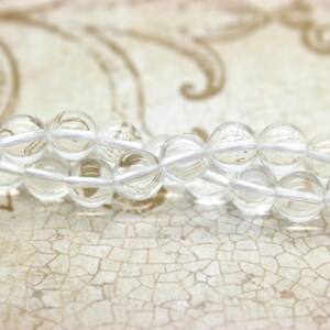May include: A strand of clear quartz beads strung on white thread. The beads are round and smooth, and they are arranged in a loose, flowing pattern.