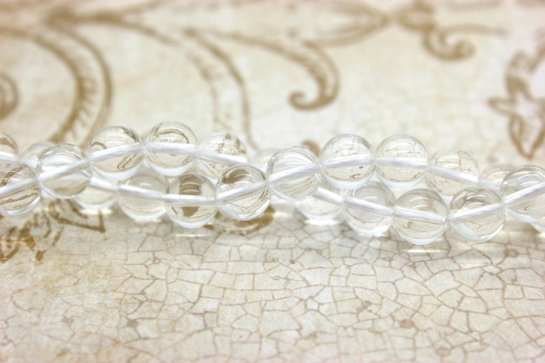 May include: A strand of clear quartz beads strung on white thread. The beads are round and smooth, and they are arranged in a loose, flowing pattern.