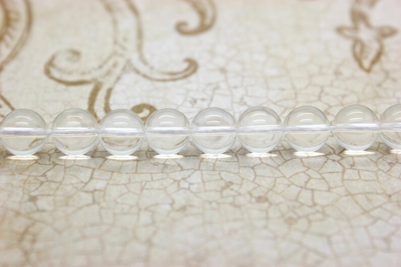 May include: A strand of clear glass beads, strung together on a white thread. The beads are round and smooth, and they reflect the light beautifully.