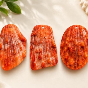 May include: Three orange-red shell pendants with a textured surface and a hole at the top for stringing. The pendants are in different shapes: irregular, rectangular, and oval. The background is a soft, off-white color.