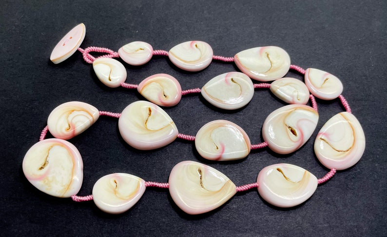 Puede incluir: Una hebra de cuentas de concha rosa y blanca con un cord&oacute;n rosa. Las cuentas tienen forma de l&aacute;grima y tienen un patr&oacute;n de remolino.