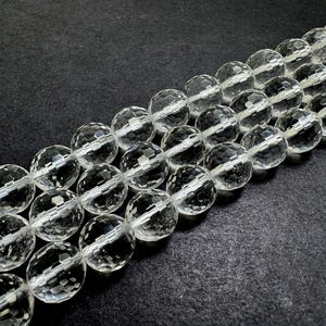 May include: Strands of clear, faceted crystal beads. The beads are round with multiple facets, creating a sparkling effect. The beads are strung together, and the background is a dark, textured surface. The beads are a neutral color and could be used for jewelry making.