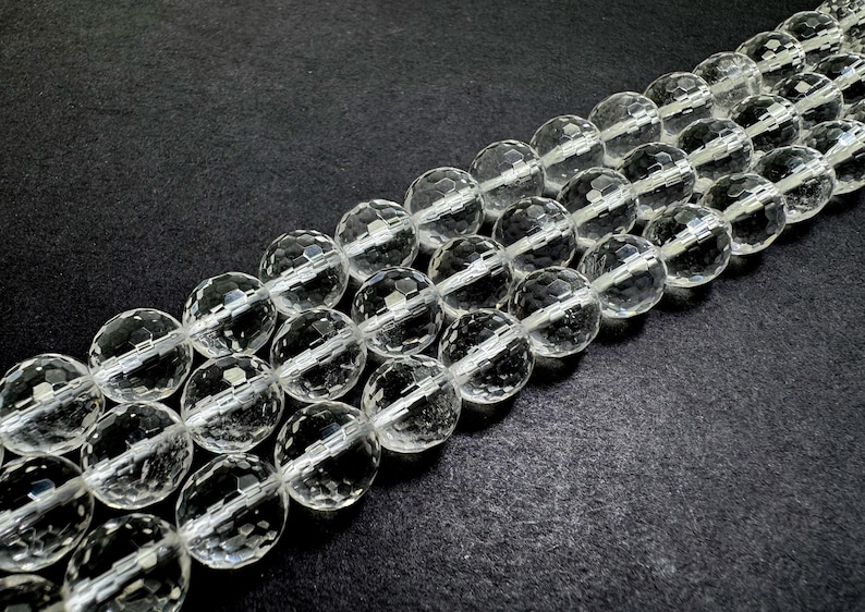 May include: Strands of clear, faceted crystal beads. The beads are round with multiple facets, creating a sparkling effect. The beads are strung together, and the background is a dark, textured surface. The beads are a neutral color and could be used for jewelry making.