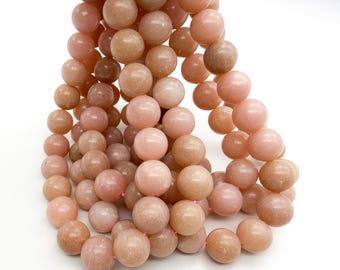 May include: A strand of round, smooth, peach-colored gemstone beads. The beads exhibit a subtle, semi-translucent quality with variations in tone ranging from light pink to soft orange, strung together on a thin, bright pink cord.