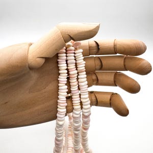 Op de afbeelding: Een ketting van schelpkralen in roze en witte tinten. De platte, schijfvormige kralen zijn aan elkaar geregen tot een ketting. De ketting wordt vastgehouden door een houten hand tegen een witte achtergrond. De kralen hebben een natuurlijke, organische uitstraling.