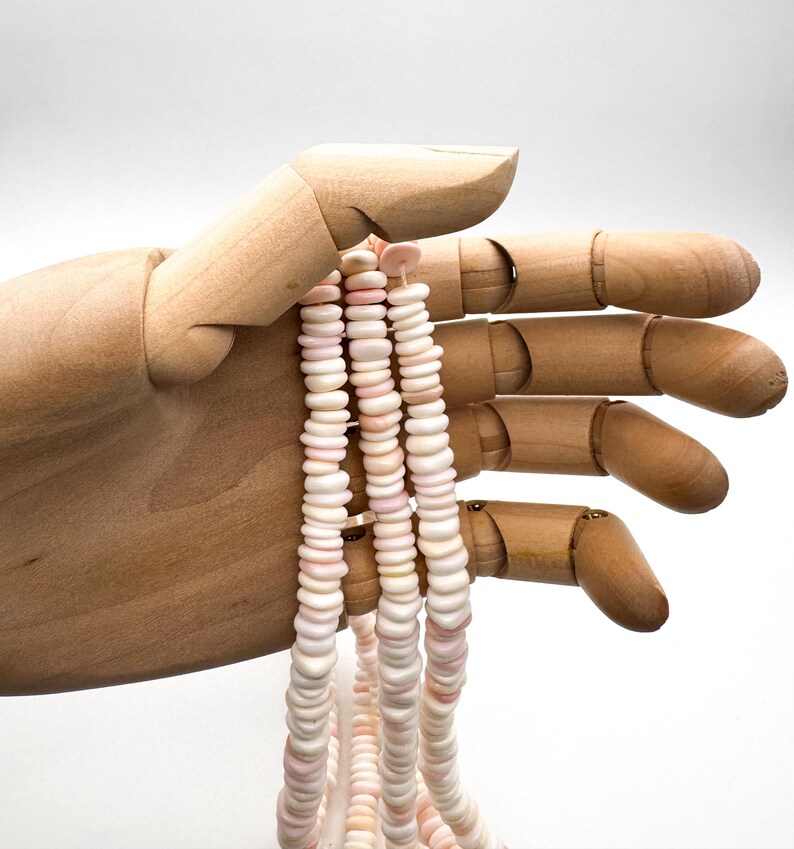 Op de afbeelding: Een ketting van schelpkralen in roze en witte tinten. De platte, schijfvormige kralen zijn aan elkaar geregen tot een ketting. De ketting wordt vastgehouden door een houten hand tegen een witte achtergrond. De kralen hebben een natuurlijke, organische uitstraling.