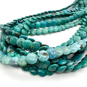 May include: A collection of multiple strands of turquoise beads, each featuring a flat, circular coin shape. The beads display a range of blue and green hues with natural dark matrix veining, arranged in overlapping layers to show their texture.