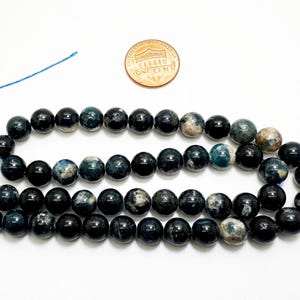 May include: Three strands of dark blue and black gemstone beads. The beads are round and polished. A one-cent coin is next to the beads for size comparison. The beads are strung on a blue string.