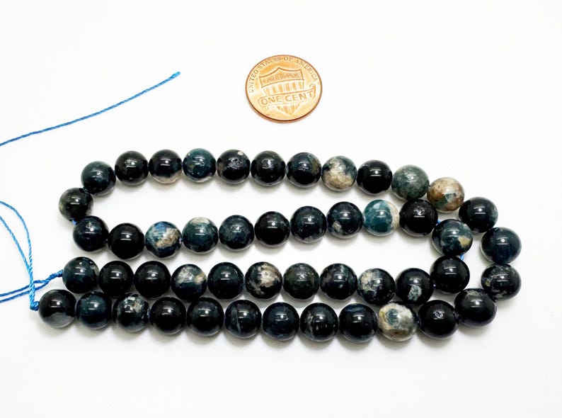 May include: Three strands of dark blue and black gemstone beads. The beads are round and polished. A one-cent coin is next to the beads for size comparison. The beads are strung on a blue string.