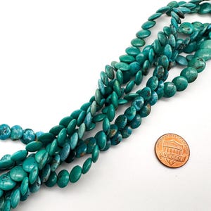 May include: A strand of round, flat turquoise beads with natural dark brown matrix veining. The beads are arranged in a loose, overlapping cluster. A copper-colored one-cent coin is placed next to the beads to provide a sense of scale.