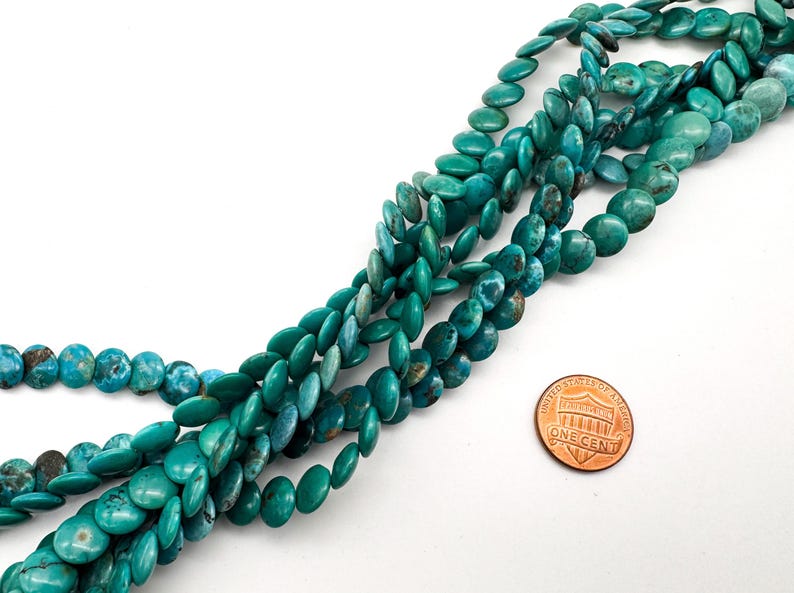 May include: A strand of round, flat turquoise beads with natural dark brown matrix veining. The beads are arranged in a loose, overlapping cluster. A copper-colored one-cent coin is placed next to the beads to provide a sense of scale.