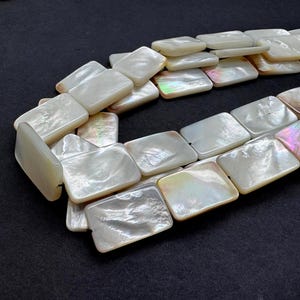 May include: A necklace composed of rectangular, iridescent mother-of-pearl beads. The beads are a creamy white color with hints of pink, green, and gold. The necklace is displayed on a dark background, highlighting the natural luster of the beads.