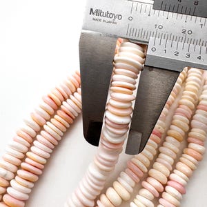 Op de afbeelding: Close-up van strengen roze en witte schelpkralen die worden gemeten met een Mitutoyo schuifmaat. De kralen zijn schijfvormig en hebben een natuurlijk, organisch uiterlijk. De schuifmaat toont metingen in millimeters.