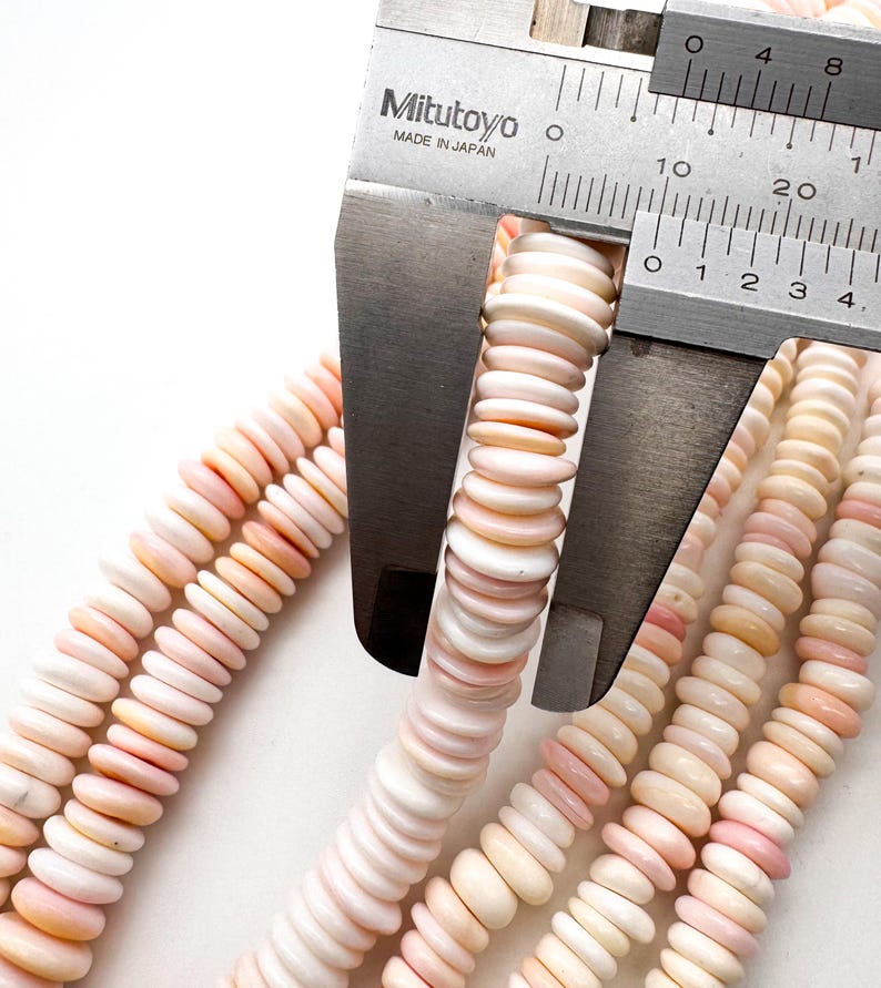 Op de afbeelding: Close-up van strengen roze en witte schelpkralen die worden gemeten met een Mitutoyo schuifmaat. De kralen zijn schijfvormig en hebben een natuurlijk, organisch uiterlijk. De schuifmaat toont metingen in millimeters.