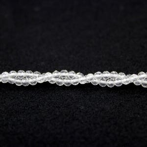 May include: A close-up of a strand of clear, faceted beads. The beads are small and round, strung together to form a delicate necklace or bracelet. The background is a solid black, highlighting the transparency of the beads.