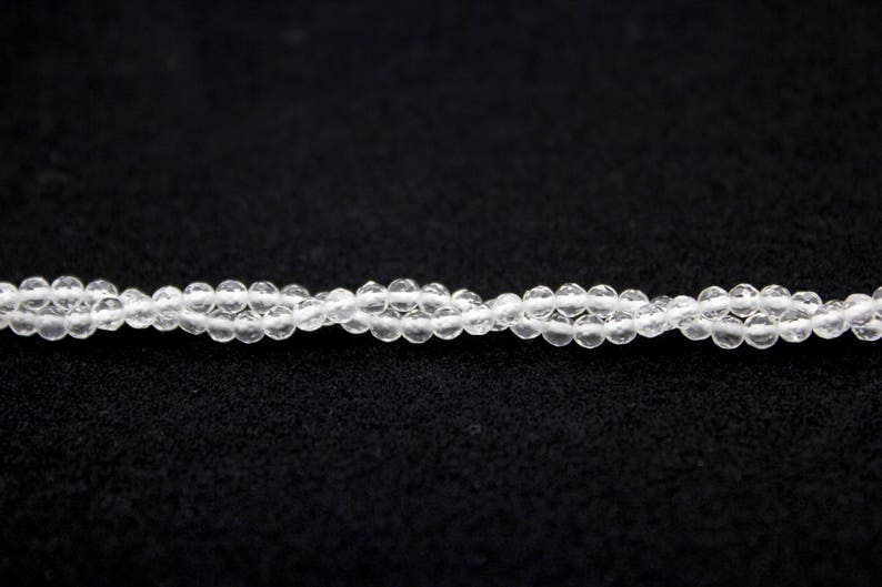May include: A close-up of a strand of clear, faceted beads. The beads are small and round, strung together to form a delicate necklace or bracelet. The background is a solid black, highlighting the transparency of the beads.