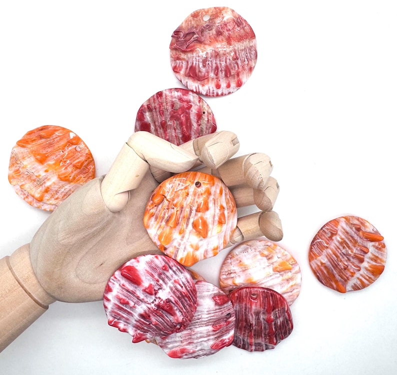 May include: A collection of colorful, round seashells in shades of orange, red, and white. The shells have a textured surface and a small hole at the top. The shells are scattered on a white background.