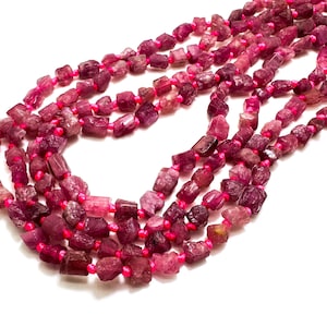 May include: Strands of rough-cut, deep pink tourmaline beads. The beads are strung on bright pink cord, creating a layered effect. The gemstones vary in size and shape, with a natural, unpolished appearance.