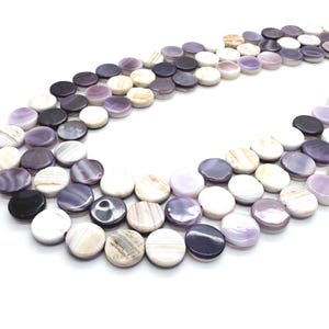 May include: A collection of flat, round beads in shades of purple, white, and black. The beads are arranged in a flowing pattern against a white background. The beads have a polished, smooth surface, and are likely used for jewelry making.
