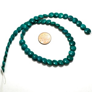 May include: A strand of round, flat, teal-colored beads arranged in a circular shape. A copper-colored United States penny is placed in the center of the bead circle to provide a visual reference for the size of the individual beads.