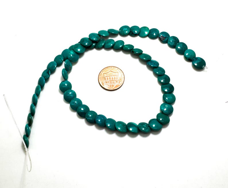 May include: A strand of round, flat, teal-colored beads arranged in a circular shape. A copper-colored United States penny is placed in the center of the bead circle to provide a visual reference for the size of the individual beads.