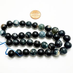 May include: A collection of dark blue and black gemstone beads, with green and grey variations. A penny is included for scale. The beads are strung on blue thread, suitable for crafting jewellery.