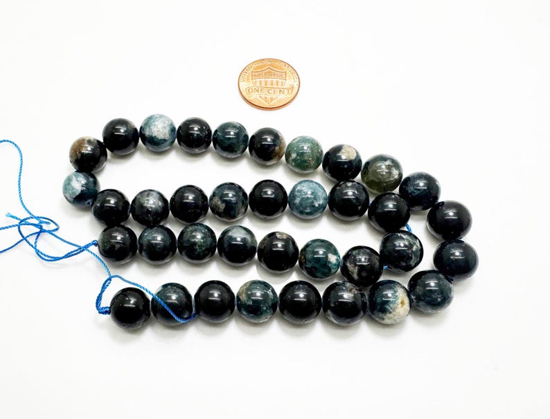 May include: A collection of dark blue and black gemstone beads, with green and grey variations. A penny is included for scale. The beads are strung on blue thread, suitable for crafting jewellery.