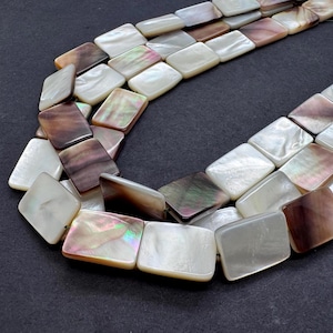 Puede incluir: Hebras de cuentas rectangulares de nácar en tonos blancos, marrones e iridiscentes. Las cuentas están ensartadas, creando un diseño de collar o pulsera. Las cuentas tienen una superficie lisa y pulida.