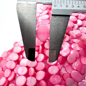 May include: Close-up of pink oval beads with a caliper measuring one bead. The beads are a light pink color and have a smooth, polished surface. The caliper is silver and has black markings. The image shows a pile of the beads.