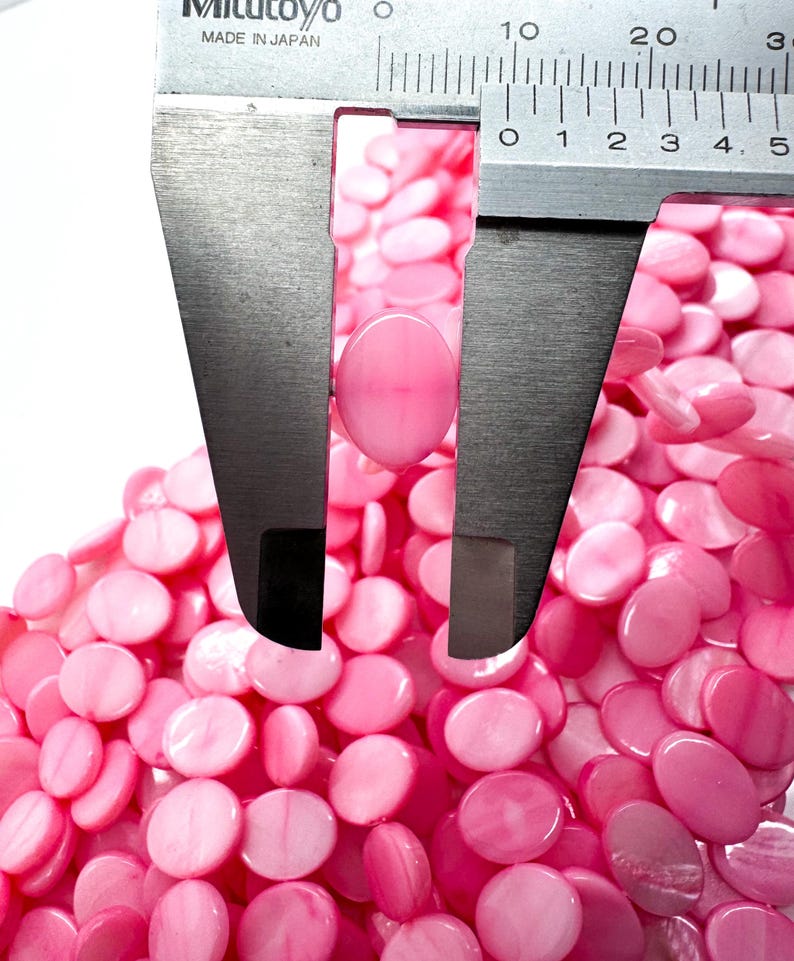 May include: Close-up of pink oval beads with a caliper measuring one bead. The beads are a light pink color and have a smooth, polished surface. The caliper is silver and has black markings. The image shows a pile of the beads.