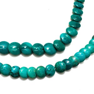 May include: A strand of round, flat turquoise beads arranged in two curved rows. The beads feature a vibrant teal-blue color with natural dark brown matrix veining throughout, creating a smooth, polished surface texture on each individual bead.