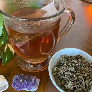 May include: A glass mug filled with a dark brown tea with a tea bag still inside. A small white bowl filled with dried herbs sits next to the mug. A small amethyst crystal and a clear crystal are on the table.