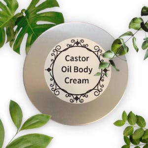 Peut inclure: Une boîte en étain argentée avec une étiquette blanche sur laquelle est inscrit "Castor Oil Body Cream" en lettres noires. L'étiquette est bordée d'un motif noir décoratif.