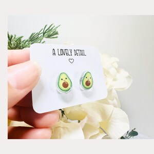 May include: A pair of avocado-shaped stud earrings with a smiling face on each avocado. The earrings are on a white card with the text "A Lovely Detail" printed on it.