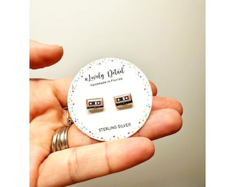 May include: A pair of sterling silver cassette tape stud earrings. The earrings are small, rectangular, and feature a detailed design of a cassette tape. The earrings are displayed on a white card with the text "a Lovely Detail" and "Handmade in Florida".