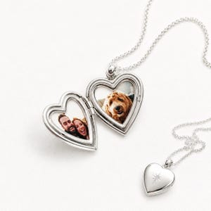 Puede incluir: Un collar con relicario en forma de corazón de plata con una cadena fina. El relicario está abierto y muestra dos fotografías en su interior: una de una pareja sonriente y otra de un perro dorado. Se ve un segundo relicario cerrado con una estrella.