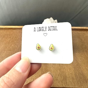May include: A pair of avocado-shaped stud earrings. Each earring features a smiling avocado design with a yellow pit and green exterior. The earrings are displayed on a white card with the text "A LOVELY DETAIL" and a heart symbol.