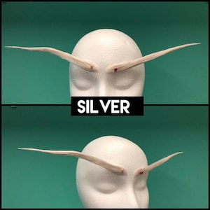 May include: A pair of long, pointed, silver-colored eyebrows made of synthetic hair fibers, displayed on a white mannequin head. Each eyebrow is secured with a small, round, colored pin at the inner corner. The text SIL VER is centered.