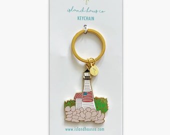 May include: Gold keychain featuring a white lighthouse with a red, white, and blue American flag on the side. The lighthouse is surrounded by gray rocks and green grass. The keychain also includes a small gold charm with a palm tree design. The text "island haus co KEYCHAIN" and "www.islandhausco.com" are printed on the card.