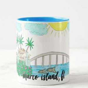 May include: A white ceramic coffee mug with a blue rim. The mug features a hand-drawn illustration of a bridge, palm trees, a sun, clouds, and dolphins. The text "marco island, fl" is written in black at the bottom of the mug.