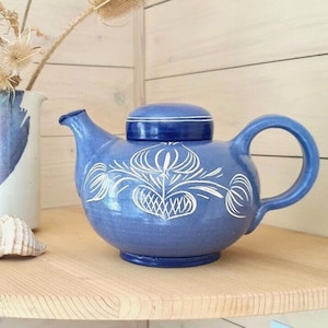 Könnte beinhalten: Eine blaue Keramik-Teekanne mit Deckel und gebogenem Griff. Die Teekanne hat ein weißes Blumendesign. Eine weiße Keramikkrug mit blauen Akzenten und eine Muschel befinden sich ebenfalls auf dem Holzregal. Getrocknete Blumen sind im Krug.