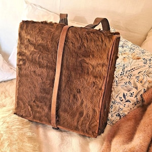 Puede incluir: Una mochila rectangular marrón con un exterior texturizado similar a la piel. La bolsa tiene un cierre de correa de cuero y dos correas de cuero para los hombros. La mochila está sobre almohadas y una manta.