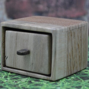 Puede incluir: Una pequeña caja de madera con un cajón. La caja está hecha de madera clara con un acabado natural. El cajón tiene una pequeña perilla de madera.
