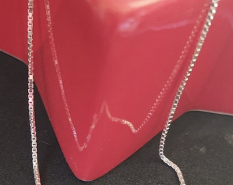 May include: A silver chain necklace with a lobster clasp. The chain is box style and is lying on a red surface.