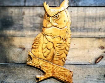 May include: A wooden carving of an owl with detailed feathers and a branch. The owl is carved in a light brown color and has a realistic appearance.